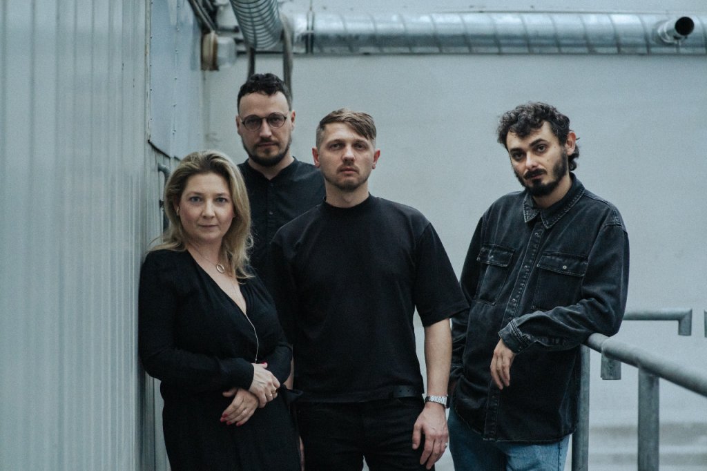 Zdjęcie przedstawia Martę Czyż i członków kolektywu Open Group. Od lewej: Yuriy Biley, Pavlo Kovach, Anton Varga. Osoby są ubrane na czarno i kontrastują z białym, industrialnym tłem. 