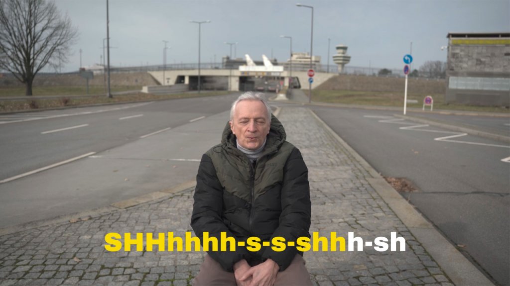Zdjęcie przedstawia kadr z wideo Powtarzajcie za mną. Starszy mężczyzna siedzi samotnie pośrodku ulicy z zamkniętymi oczami.