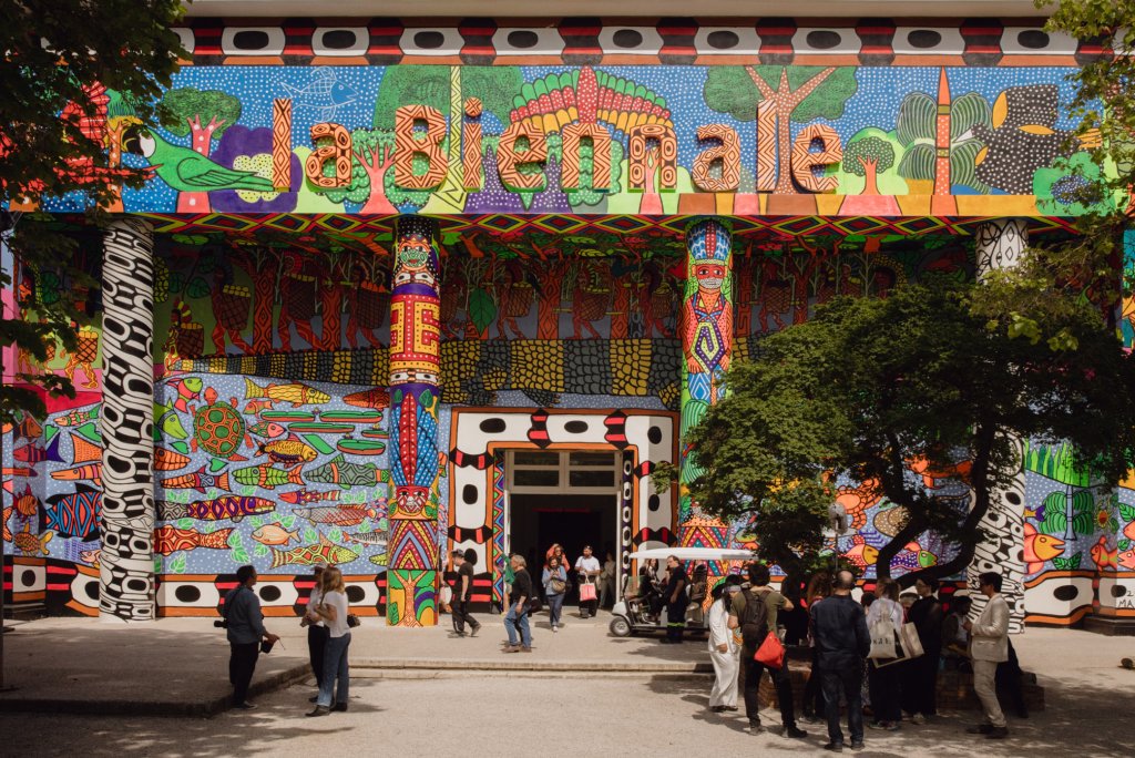 Zdjęcie przedstawia pokrywający budynek z kolumnami kolorowy, mozaikowy mural nawiązujący do rdzennych mieszkańców Brazylii i Peru, stworzony przez kolektyw artystyczny MAHKU. Napis nad wejściem do budynku: la Biennale.