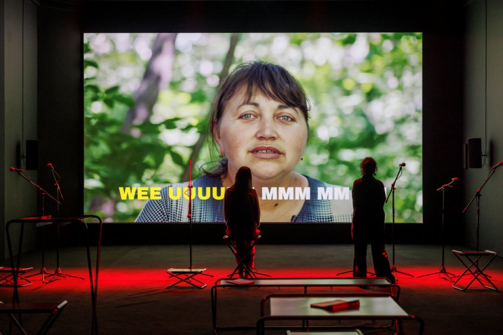 Zdjęcie przedstawia kadr z wideo Powtarzajcie za mną, przedstawiający kobietę w średnim wieku, wyświetlony na oświetlonej na czerwono scenie, na której rozmieszczono mikrofony na statywach. Stoją przy nich performerzy z kolektywu Open Group.