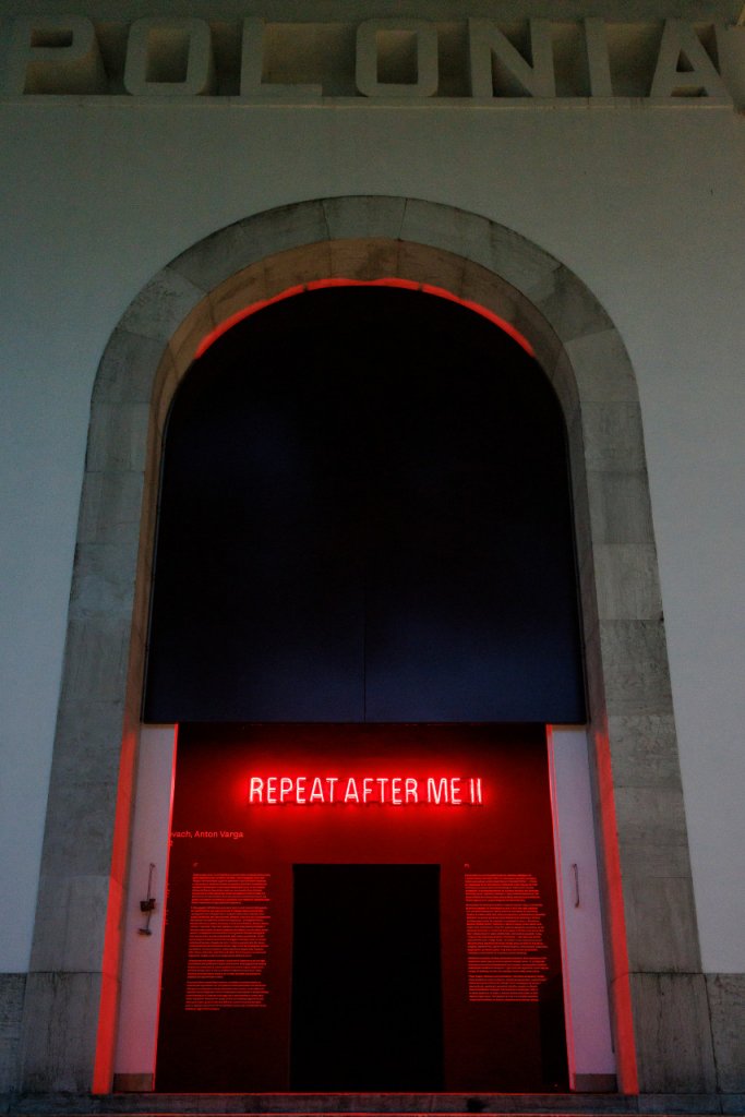 Zdjęcie przedstawia widok na wystawę w Pawilonie Polski na Biennale. W bramie umieszczono czerwony neon z napisem Powtarzajcie za mną 2.