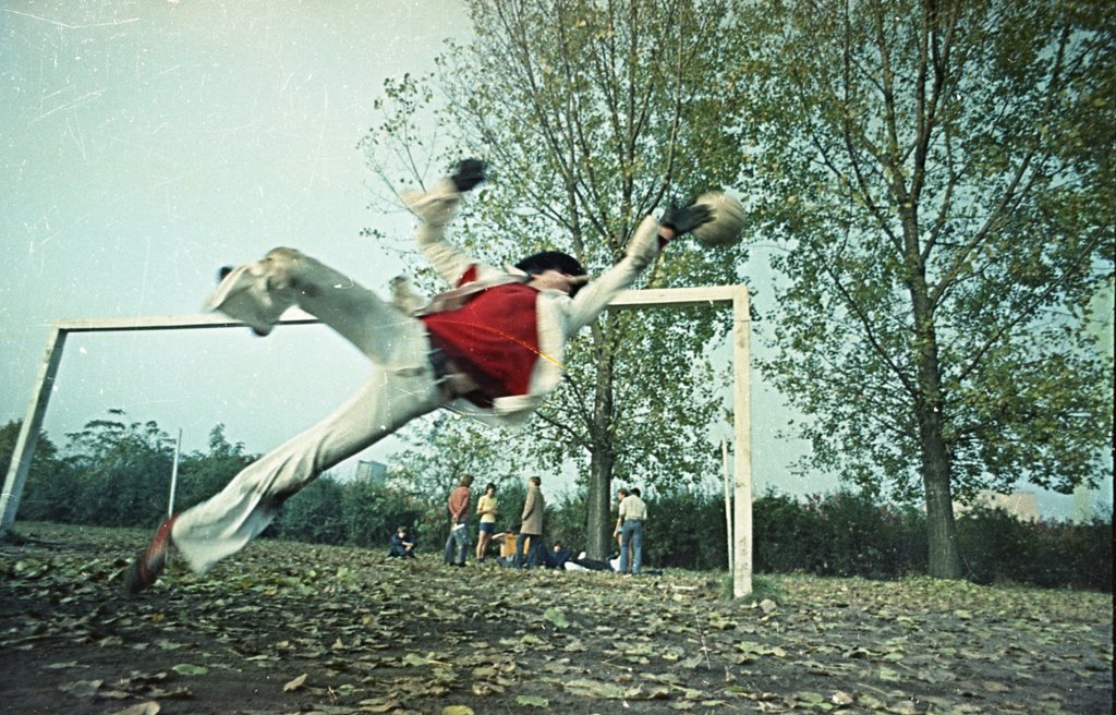 Kadr z filmu Goalkeeper, 1975–1977. Na zdjęciu widzimy artystę w dynamicznej pozie rzutu bramkarskiego, uchwyconego w locie, tuż przed spektakularną obroną. Ubrany w elegancki biały garnitur, czerwony bezrękawnik i kapelusz, Sosnowski kontrastuje z prowizorycznym boiskiem i jesiennym pejzażem.