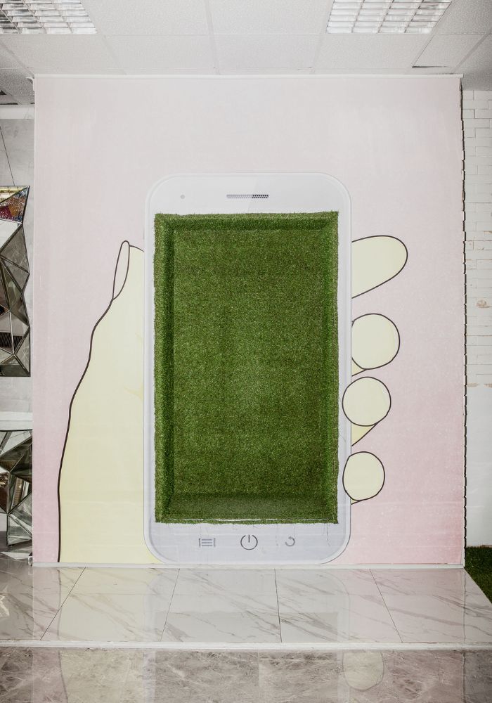 Instalacja artystyczna przedstawiająca rysunek dłoni trzymającej smartfon, którego ekran wypełnia zielona sztuczna trawa.