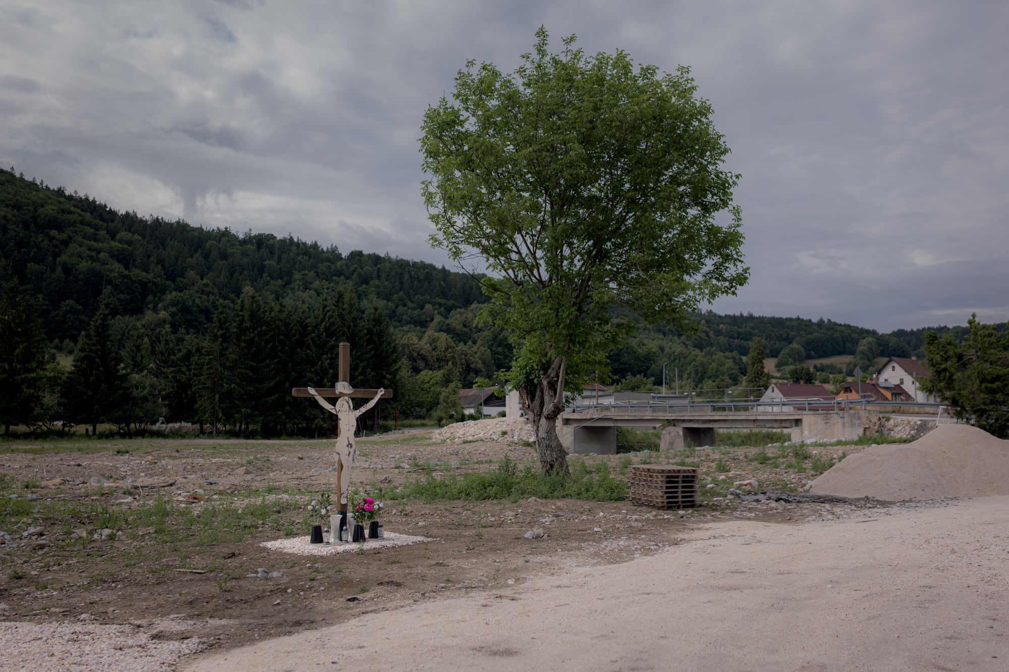 Radochów rok po powodzi (2025). Krzyż z figurą Chrystusa stoi przy drodze, obok ustawione kwiaty i znicze. W tle pojedyncze domy, mostek i zielone wzgórza.