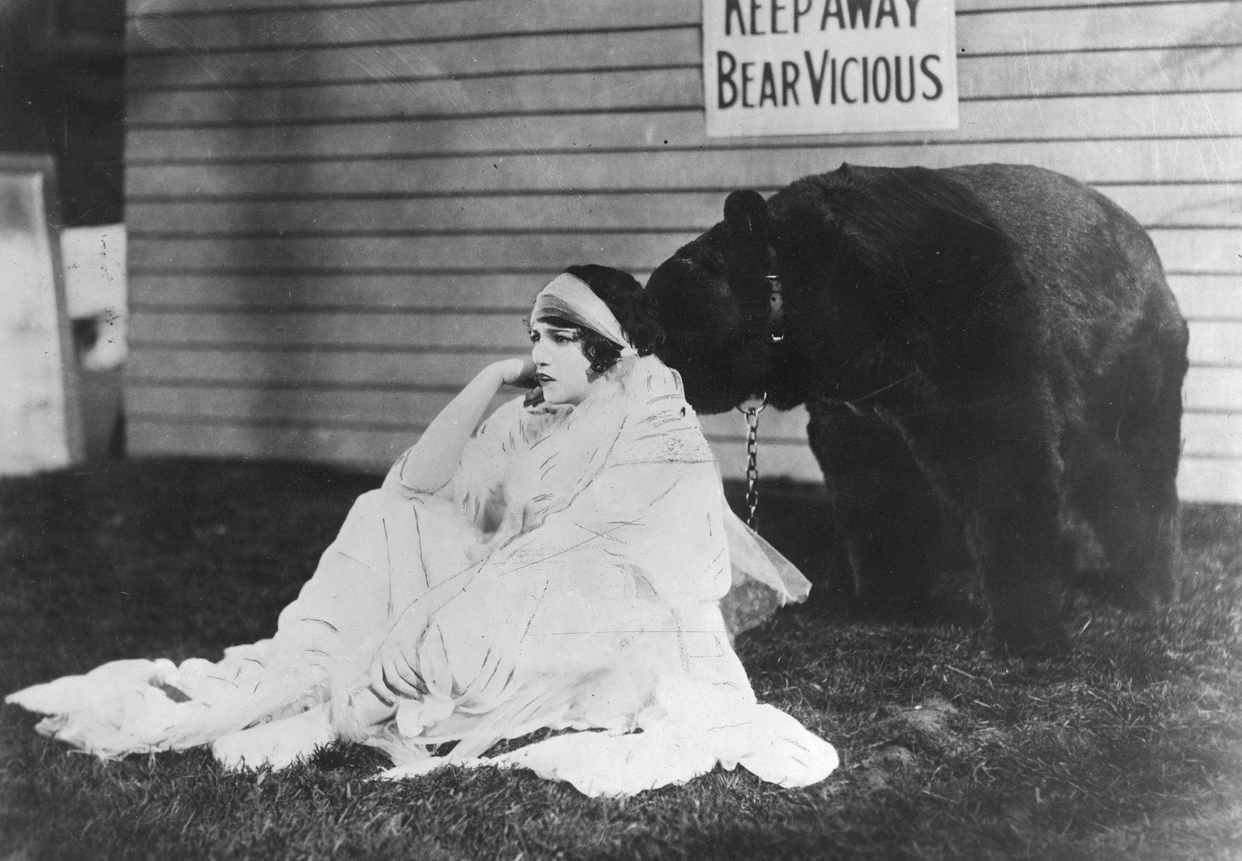 Czarno-biała fotografia archiwalna przedstawia młodą kobietę siedzącą na trawie. Ubrana jest w jasną, zwiewną suknię i opiera głowę na dłoni. Ma krótkie, ciemne włosy przewiązane opaską. Za nią widoczna jest sylwetka niedżwiedzia z łańcuchem przy uprzęży. Zdjęcie: Narodowe Archiwum Cyfrowe, domena publiczna.
