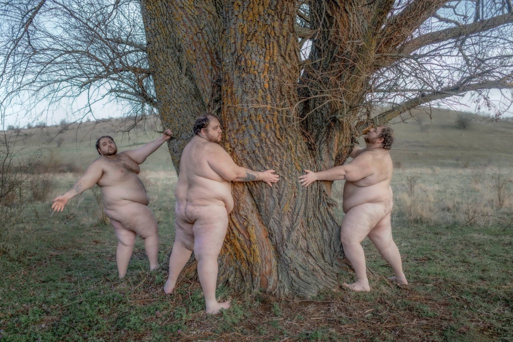Fotografia Artema Humilevskiego: naga, korpulentna postać mężczyzny pokazana dwukrotnie w jednym kadrze stoi przy dużym drzewie na łące. Jedna z postaci obejmuje pień drzewa, druga stoi obok z rozłożonymi rękami. W tle bezlistne gałęzie i otwarty krajobraz.